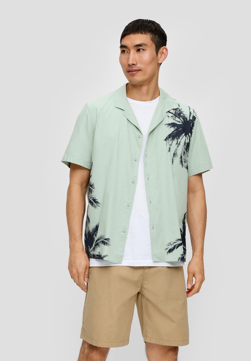 Lichtgroen overhemd met korte mouwen en zwart palmboomprint aan één kant, met knoopsluiting aan de voorkant, gecombineerd met een wit T-shirt en kaki shorts.