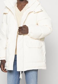 Bílá péřovka s kapucí, elastickými manžetami a předními kapsami. Obsahuje zipový uzávěr a jemnou texturu, nošena přes béžový svetr.