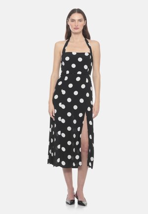 Donna che indossa un vestito midi nero con collo halter, pois bianchi e spacco alto sulla coscia, abbinato a tacchi neri a punta.