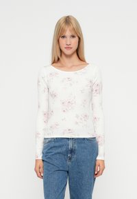 Langarmoberteil aus weißem Rippmaterial mit pinkem Blumenmuster. Es verfügt über einen weiten Ausschnitt und eine taillierte Passform, kombiniert mit hochgeschnittenen Jeans.