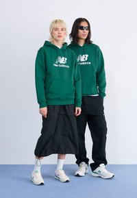 Sweatshirts à capuche verts avec un logo "new balance" blanc ; associés à un pantalon cargo noir et des baskets blanches avec des accents verts.