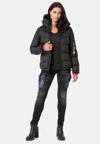 Schwarze Steppjacke mit Kapuze, mit rosa Akzenten an den Bündchen. Kombiniert mit dunklen Jeans, blauen Taschenakzenten und schwarzen Stiefeletten mit Absatz.
