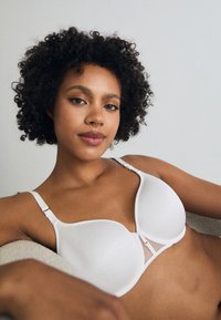 White bra with smooth fabric, contoured cups, and mesh accent at the center. Features adjustable straps and gold-toned hardware.