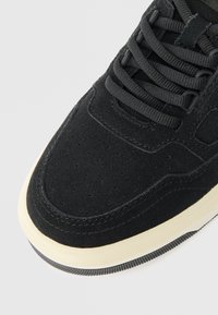 Crni nubuk tenisice s perforiranim detaljima, crne vezice i krem sredinom na bijeloj pozadini.