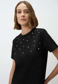 Zwarte t-shirt van zacht materiaal, met verspreide zilveren versieringen aan de voorkant. Klassieke ronde hals en korte mouwen.