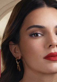 Gros plan sur un visage avec une peau lisse, des cheveux noirs, des sourcils bien définis, des lèvres rouges vives et une boucle d'oreille en or torsadée. Un tube de rouge à lèvres rose est visible.