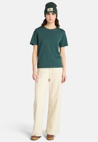 Teal T-shirt med korte ærmer og et lille logo, parret med beige brede bukser og brune sko. Derudover har det en grøn strikhue med et mærke.