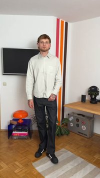 Camisa de botões cinza-claro com mangas longas, jeans escuros e sapatos esportivos pretos. O fundo apresenta paredes e móveis listrados e coloridos.