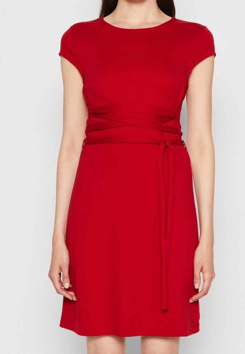 Femme portant une robe rouge à manches courtes avec une ceinture nouée autour de la taille, montrée du cou jusqu’au-dessus des genoux.