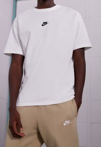 Bílý bavlněný tričko s krátkým rukávem se malým černým logem Nike na hrudi, spárované s béžovými tepláky s bílým logem.