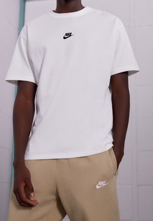 Camiseta blanca de algodón con mangas cortas que presenta un pequeño logo negro de Nike en el pecho, combinada con pantalones de chándal beige con un logo blanco.