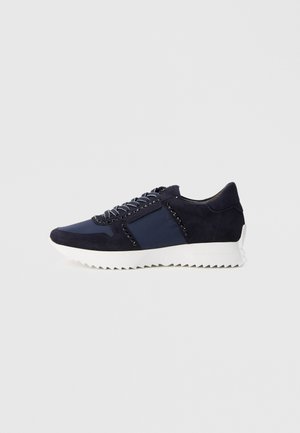 Navy blue sneaker with black stud detailing, white zigzag sole, and patterned laces on a plain light grey background.