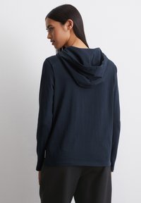 Marineblauwe hoodie van een zachte stof, met lange mouwen en een ontspannen pasvorm. Heeft een enkele achternaad en een subtiel logo-tagg aan de zijkant.