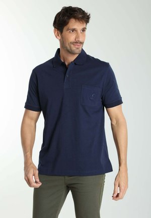 Homme portant un polo bleu marine à manches courtes avec une poche poitrine, debout devant un fond blanc, regardant vers sa gauche.