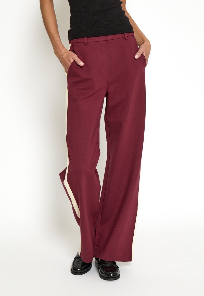 Maroon wide-leg trousers with contrasting cream side stripes, made from smooth fabric. Features a waistband, pockets, and a relaxed fit.