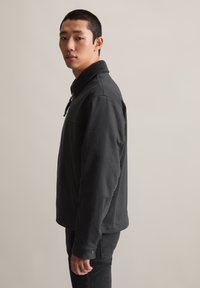 Schwarze Freizeitjacke mit Kragen, Reißverschluss und seitlichen Taschen. Der Stoff hat eine glatte Textur mit minimalen Akzenten.