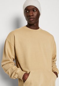 Beige katoenen sweatshirt met lange mouwen, een ronde hals en een voorknoop. De stof is soepel en vertoont een subtiele structuur.