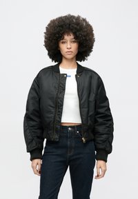 Svart bomberjakke med blank finish, ribbestrikkede mansjetter og krage. Har glidelås foran og sidelommer. Brukt over en hvit crop top.