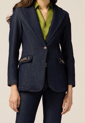 Kobieta w ciemnoniebieskim dżinsowym blezerze z kieszeniami na klapie ozdobionymi łańcuszkiem, na limezielonej koszuli i dopasowanych ciemnoniebieskich dżinsowych spodniach.