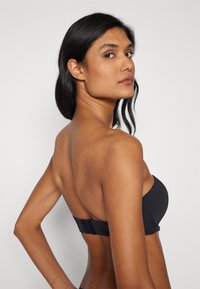 Top de bikini bandeau negro con textura suave, que cuenta con un cierre de broche en la parte posterior y un diseño minimalista.