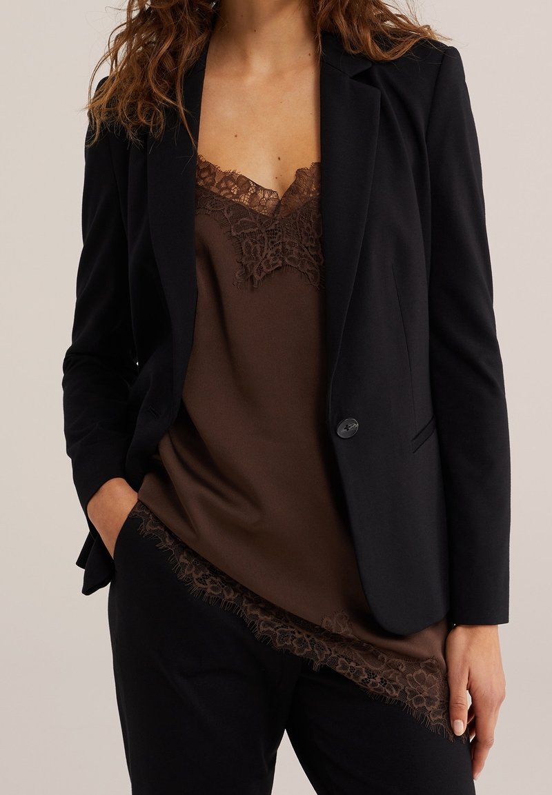 Femme portant un blazer noir sur un caraco marron à bordure en dentelle et un pantalon noir, une main dans la poche et les cheveux lâchés.