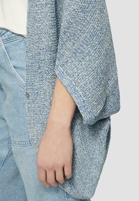 Gilet en tricot dans des tons de bleu clair et de gris, présentant une texture à mailles lâches, des épaules tombantes et une fermeture par un bouton unique.