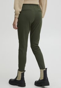 Groene slim-fit broek met een elastische tailleband en twee achterzakken, gecombineerd met zwarte enkellaarsjes met beige accenten.