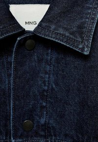 Col de veste en denim bleu foncé avec boutons noirs et étiquette de marque blanche indiquant "MNG" à l'intérieur de l'encolure.