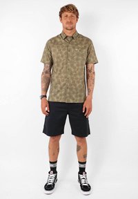 Chemise à manches courtes, verte olive, à boutons avec un motif de feuilles de palmier, associée à un short noir et des baskets montantes noires.