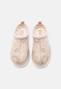Ζευγάρι μπεζ παντόφλες UGG slip-on με κεντημένα κυκλικά σχέδια και μαλακή λευκή επένδυση από μάλλινο ύφασμα, τοποθετημένες σε λευκό φόντο.