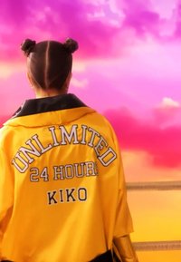 Persona con peinado de doble moño que lleva una chaqueta amarilla con el texto "UNLIMITED 24 HOURS KIKO" en la parte trasera, frente a un cielo rosa y naranja.