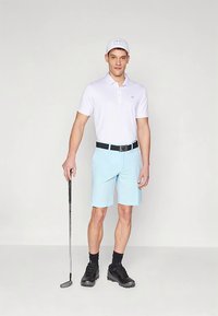 Camisa polo blanca, pantalones cortos azul claro, cinturón negro y zapatos de golf negros. Sosteniendo un palo de golf, lleva una gorra blanca con un logotipo.