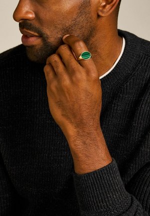 Close-up van een man die een zwarte trui draagt en zijn kin vasthoudt met een hand die is versierd met een gouden ring met een groene gestreepte steen.