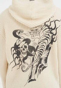 Beige geripptes Hoodie mit einem schwarzen Grafikdesign eines Schädels und eines Tigers. Der Stoff ist weich und leicht dehnbar, mit einer Kapuze.