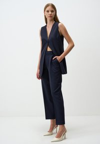 Gilet sans manches bleu marine avec un décolleté en V profond, fermeture à boutons et pantalon fuselé. Le modèle porte des chaussures à talons hauts beiges ; design et texture minimalistes.