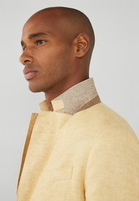 Chaqueta de lino en amarillo suave con un cuello marrón texturizado. Presenta un único bolsillo en el pecho y un detalle de botón minimalista en el cuello.