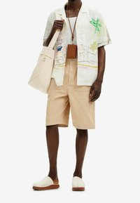 Camicia a maniche corte, con motivo di foglie e una scena di spiaggia, abbinata a pantaloni beige, una borsa leggero e zoccoli bianchi con suole marroni.