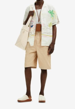 Camisa de manga corta con un estampado de hojas y una escena de playa, combinada con pantalones cortos beige, una bolsa de mano ligera y zuecos blancos con suelas marrones.