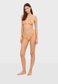 Femme debout pieds nus portant un bikini jaune à motif floral avec un haut triangle et un bas taille basse sur un fond uni clair.