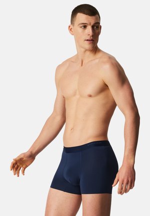 Shirtless man wearing navy blue boxer briefs, standing with arms slightly extended, looking to his left against plain light background.