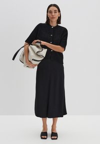 Schwarze kurzärmelige Strickjacke mit strukturiertem Strick und silbernen Knöpfen, kombiniert mit einem schwarzen Midirock und schwarzen Slip-ons, und hält eine cremefarbene Handtasche.