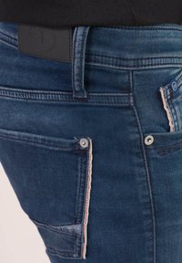 Jeans in denim blu scuro con un design aderente, caratterizzati da una toppa in pelle sul retro, dettagli in metallo sui bottoni e cuciture a contrasto sulle tasche.