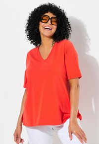T-shirt rossa a collo a V con maniche corte, tessuto morbido, vestibilità rilassata, abbinata a pantaloni bianchi e occhiali da sole oversize. Capelli ricci presenti.