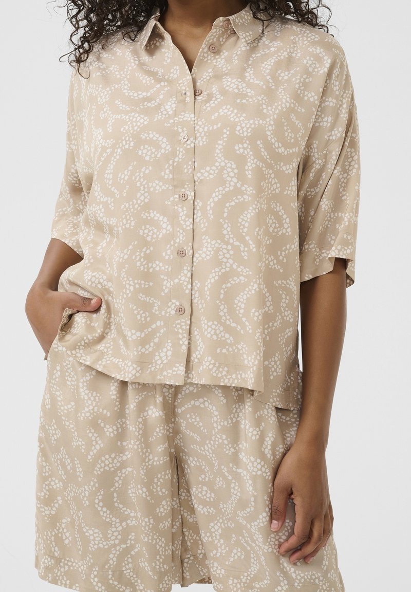 Chemise beige à manches courtes boutonnée et short assorti avec motif de points blancs irréguliers, porté par une personne avec une main dans la poche.