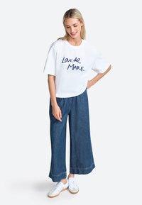 Weißes, kurzes T-Shirt mit blauen handgeschriebenen Text "Loved de Mare", kombiniert mit weit geschnittenen dunklen Jeans und weißen Sneakers.