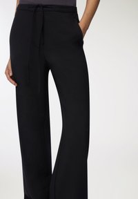 Czarne szerokie spodnie o luźnym kroju z paskiem na sznurki, gładką fakturą i bocznymi kieszeniami, pokazane od pasa w dół.