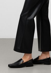 Scarpe slip-on nere in pelle con suola piatta, caratterizzate da un accento dorato e una trama liscia. Indossate con pantaloni larghi neri in ecopelle.