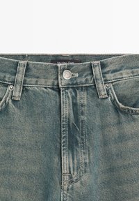 Denim jeans in light blue with a faded appearance. Features include a button closure, belt loops, and visible stitching detail.