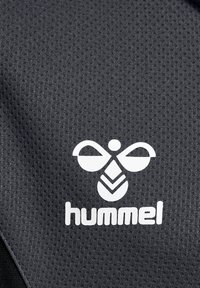 Sort tekstureret stof med et hvidt Hummel-logo med et karakteristisk bi-lignende design og brandnavn nedenfor.