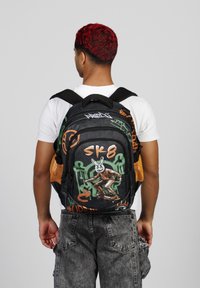 Mochila negra con diseño de graffiti en naranja y verde, que presenta una figura de patinaje. Tejido texturizado, correas acolchadas y bolsillos laterales de malla.